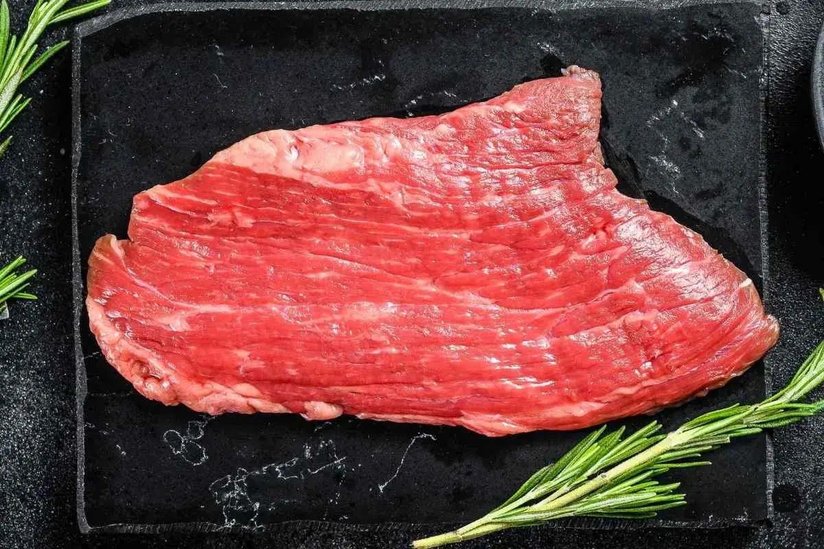 What Is Flank Steak Called at the Grocery Store? 1 Raw flank steak on a wooden cutting board, with visible grain and a textured gray countertop background