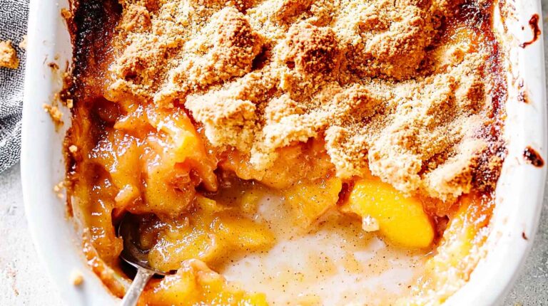 Peach cobbler in a white baking dish with golden crust and wooden spoon